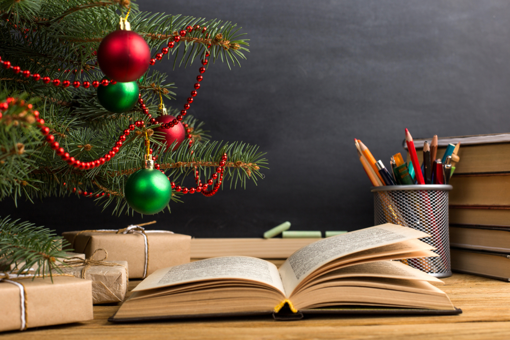 Presents under a Christmas tree, with pencils in a metal cylinder, books piled up, with a chalkboard behind the scene